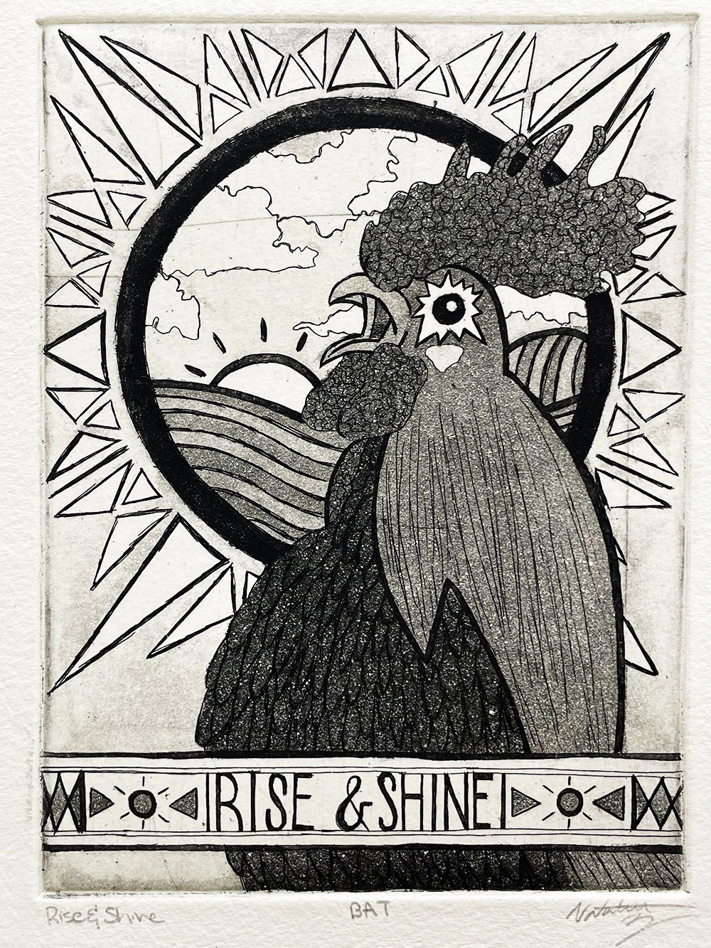 A black and white printed image composed of a rooster with the sun rising above a field behind the animal. With the words "Rise and shine" written across the bottom of the print. 
