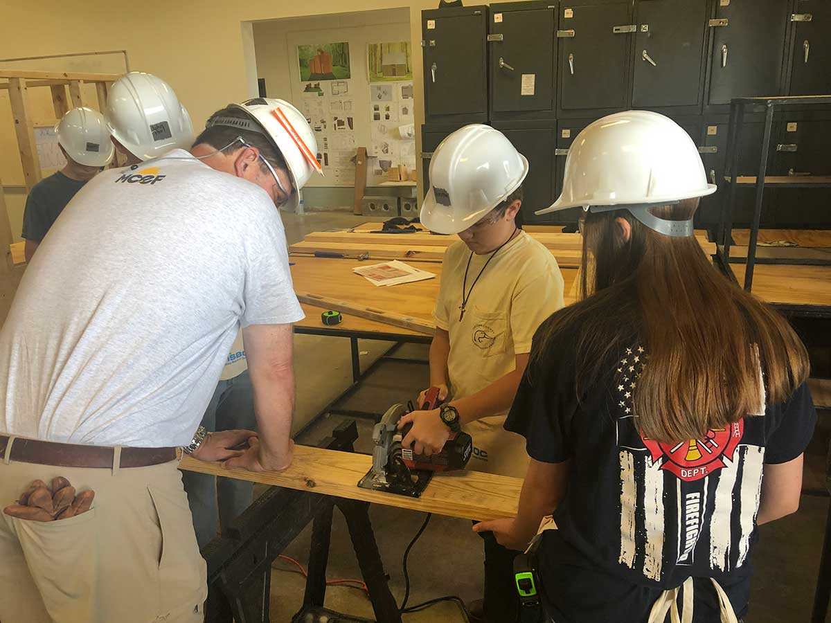student cutting wood with circular saw with help of construction professional - teammates look on