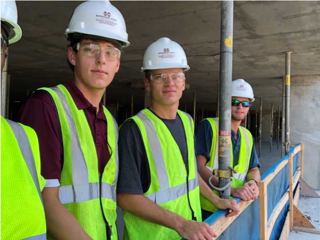 3 Building Construction Science students wearing hard hats smile for camera