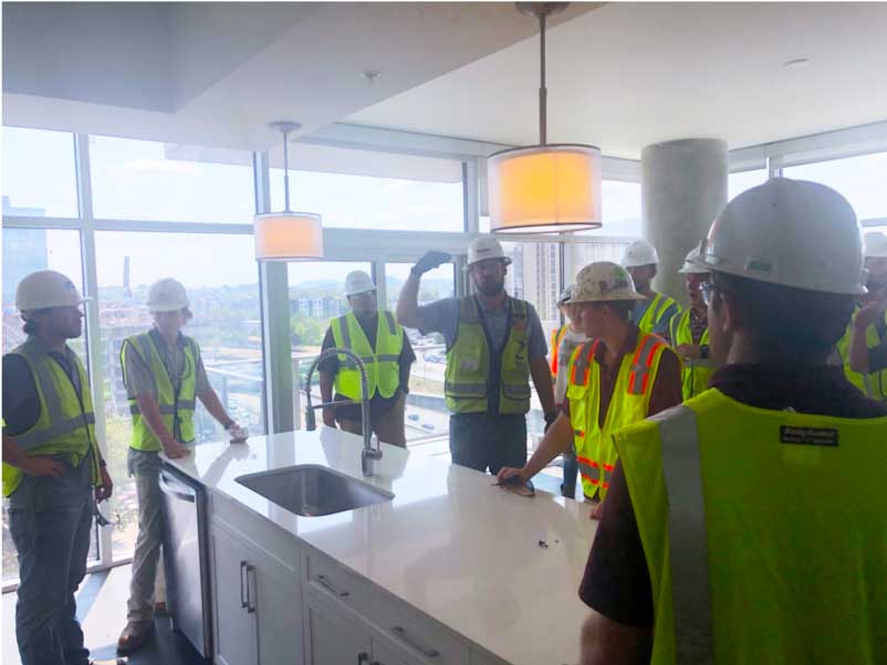 Building Construction Science students wearing safety vests and hard hats listen in group to Turner Construction reps on job site - looks like a finished kitchen