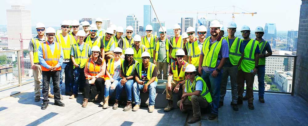 Building Construction Science students wearing safety vests and hard hats pose as a group on a field trip with a view of Nashville