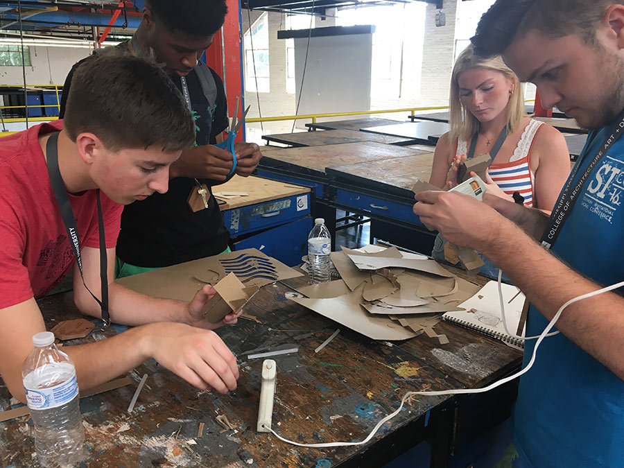 four campers use a hot glue gun and cardboard for one of their assignments. They are gathered in a circle around a studio desk