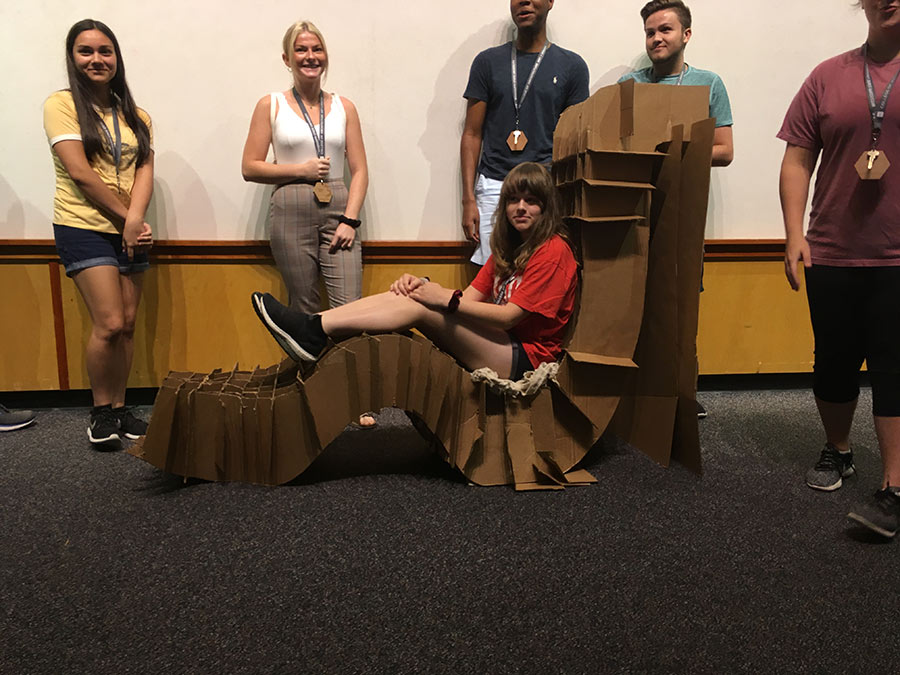 a camper sits on her group&#039;s lounge device they designed out of cardboard. group members stand around and behind her.