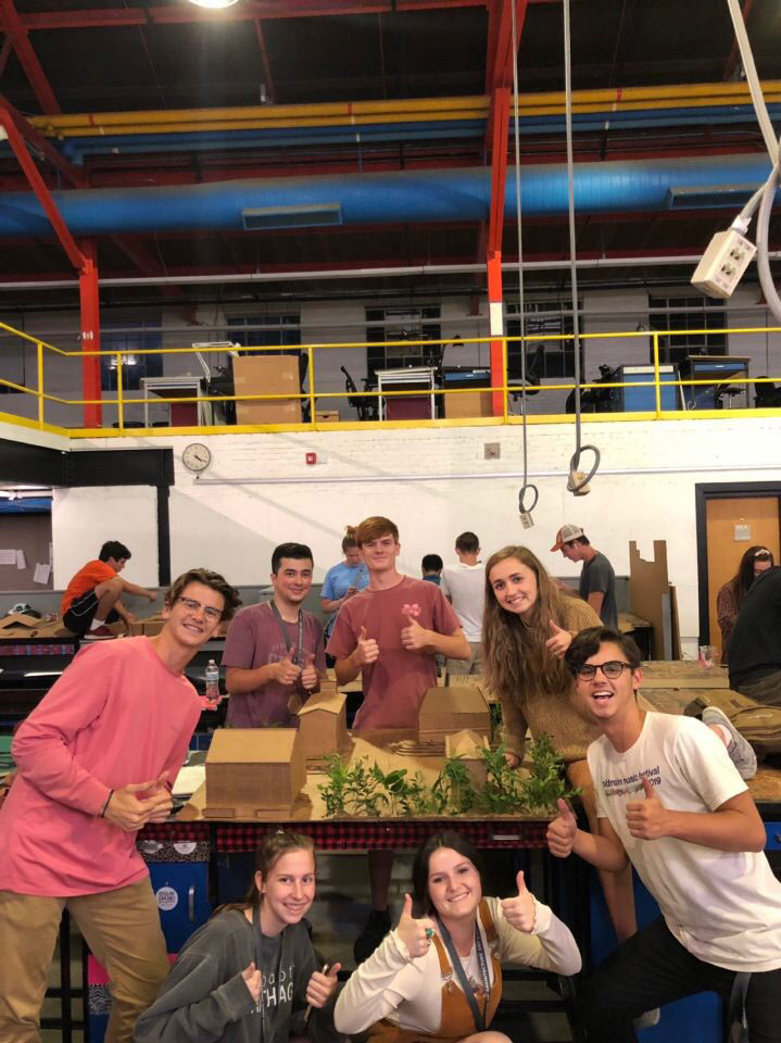 a group of campers poses with thumbs up around their project (houses made of cardboard and greenery). The project sits on a desk in the BARN area of Giles Hall.