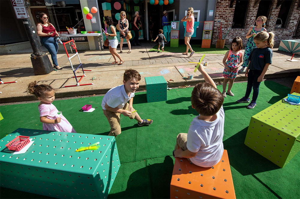 children sit on colorful seating and play on green turf while parents and others watch from sidewalk