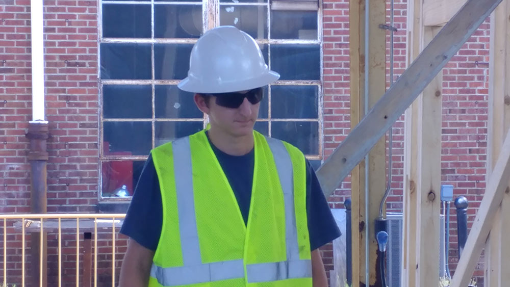 student in construction PPE and sunglasses stands outside Howell Building 