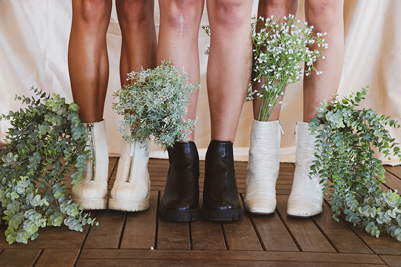 Photograph of three pairs of legs wearing boots standing on a wooden floor with plants.
