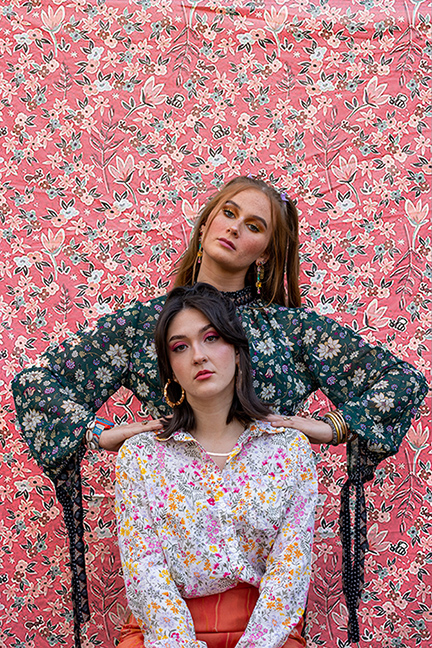 Photograph of two young women with colorful blouses standing in front of a floral background.