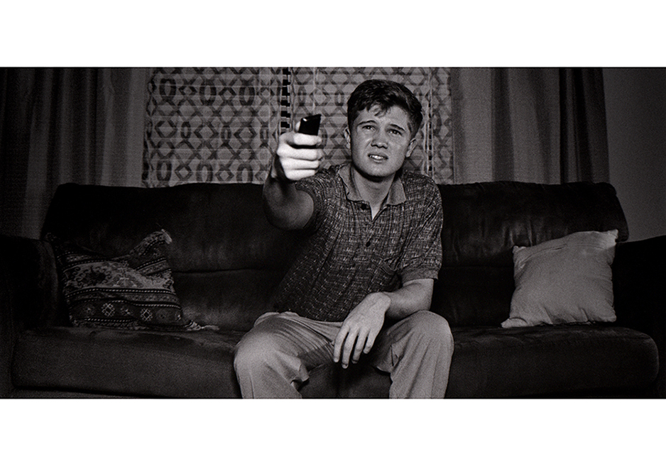 Black and white still image from a film. Image is a man sitting on a couch.