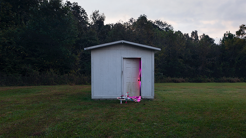 Photograph of a white shed in a grassy outdoor area. Pink light is glowing from the inside of the open door.