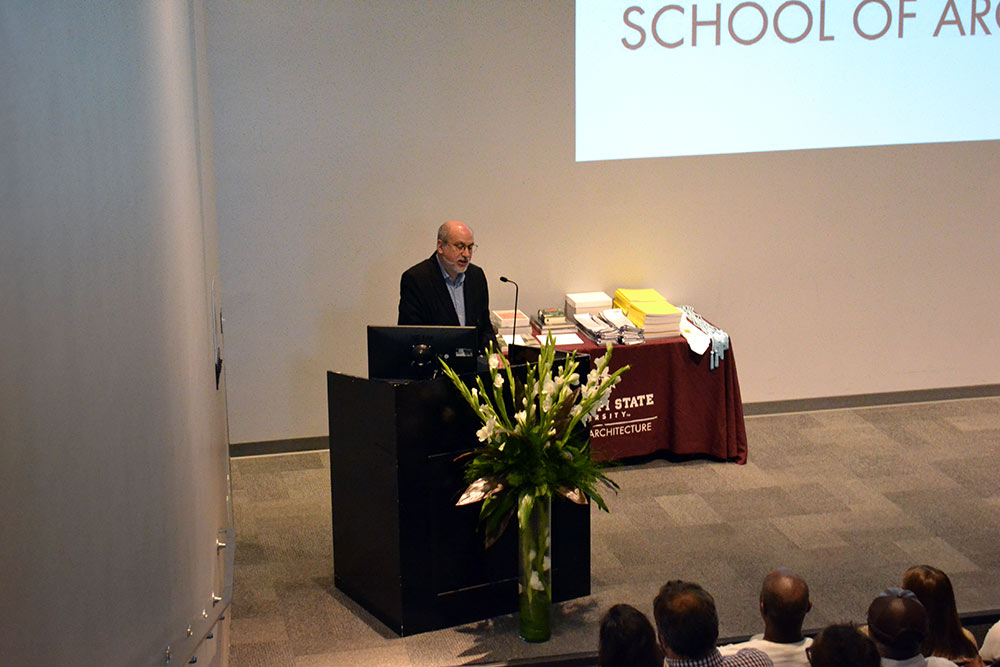 Professor John Poros at podium before audience in the Robert and Freda Harrison Auditorium in Giles Hall