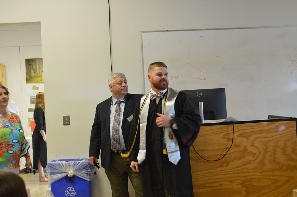 A student in his graduation robe stands with a man in a suit.