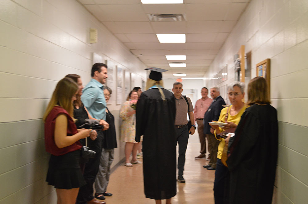 A group of people stand in a hallway, one student is wearing a graduation robe.