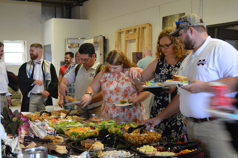 A large group of people make a plate of food from a large table holding a spread of dishes.