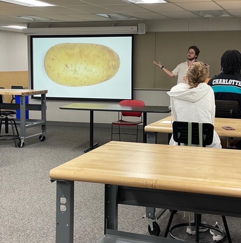 A large projector shows an image of a potato while a camper discusses something.