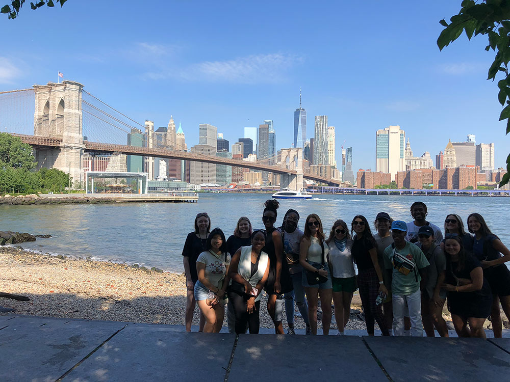 students pose with NYC skyscrapers in background and body of water, bridge