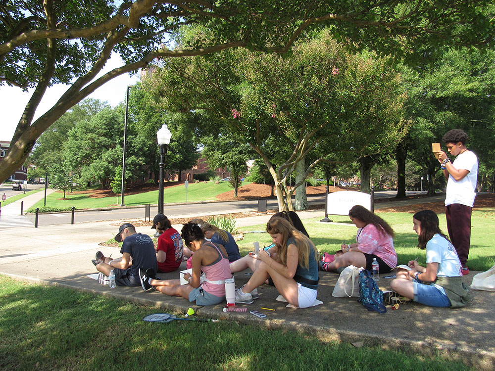 Design Discovery campers sketch in their notebooks, sitting on the ground outside.