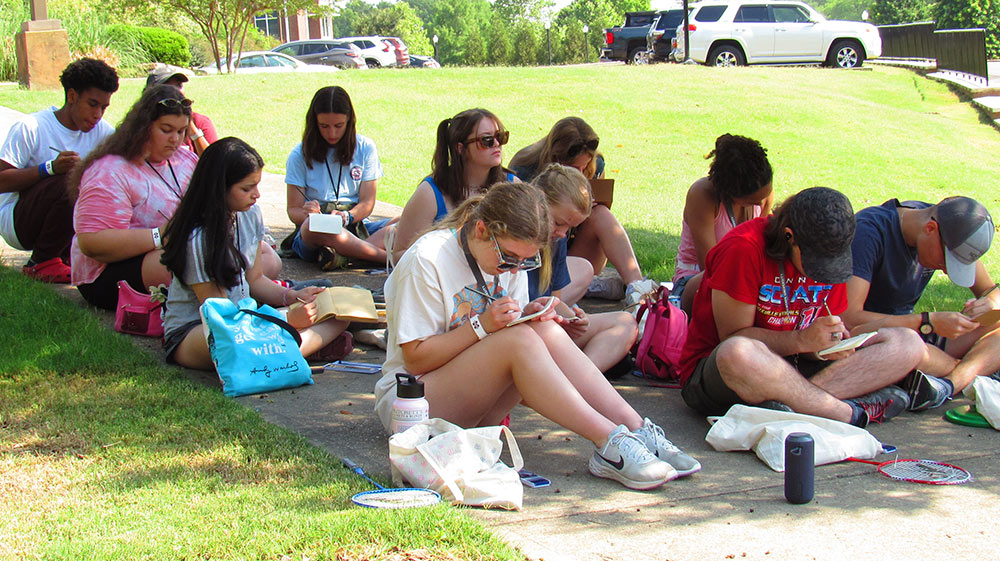 Design Discovery campers sketch in their notebooks, sitting on the ground outside.