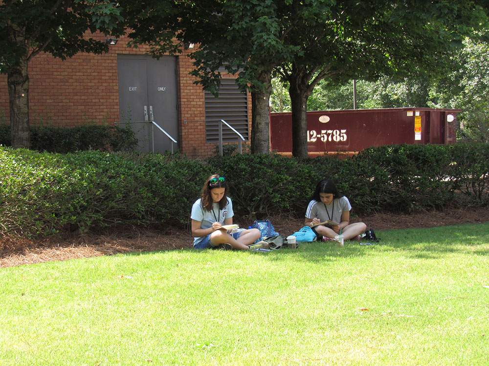 Design Discovery campers sketching in their notebooks in the grass outside.
