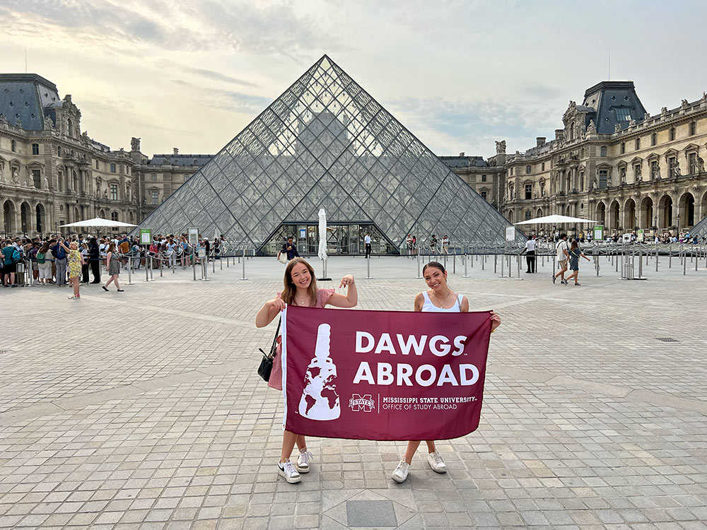 Hallie Pitts and Mackenzie Sherrell outside the Musée du Louvre in Paris, France, hold maroon DAWGS ABROAD flag