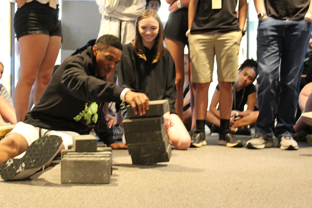 Group of Design Discovery campers observing as one camper places multiple bricks on a small object, and they start to fall.