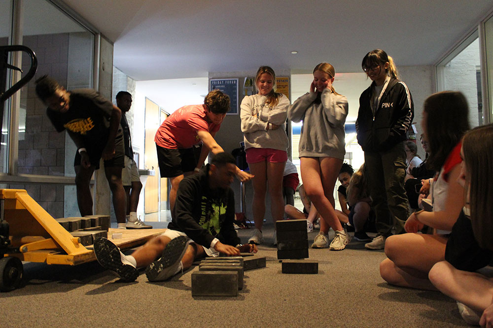 Group of Design Discovery campers observing as one camper places multiple bricks on a small object, and they fall over.