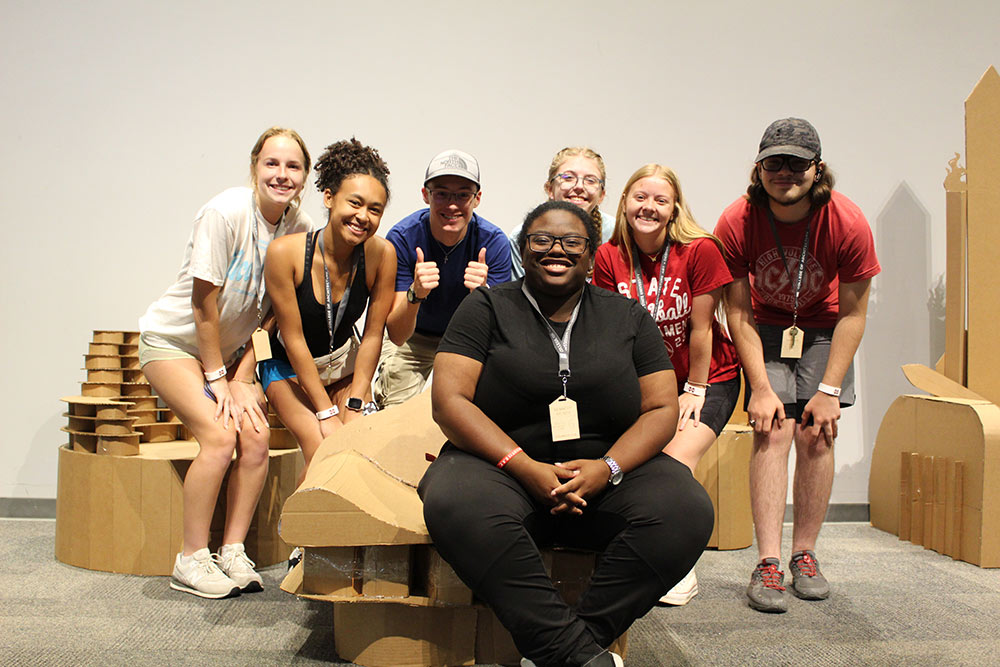 Group of campers pose around one camper who is sitting on their cardboard chair project.