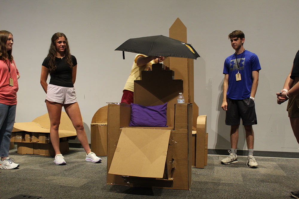 Group of campers stand around their cardboard chair project, and one camper puts an umbrella on top.