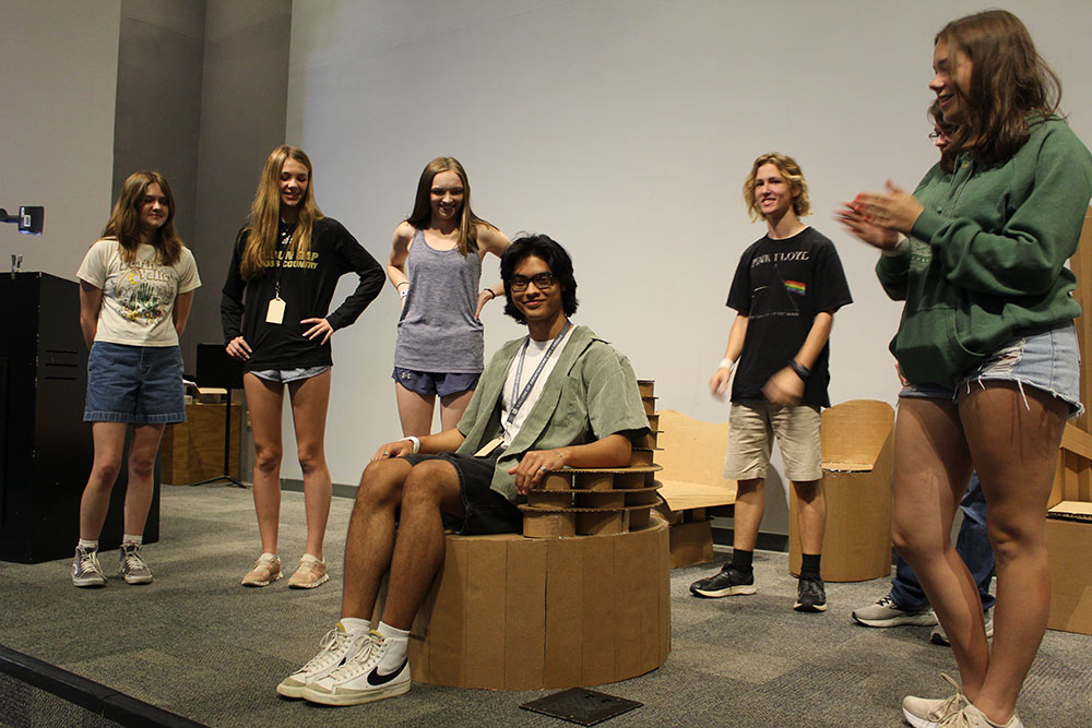 Group of campers stand around their cardboard chair project as one camper sits in it.