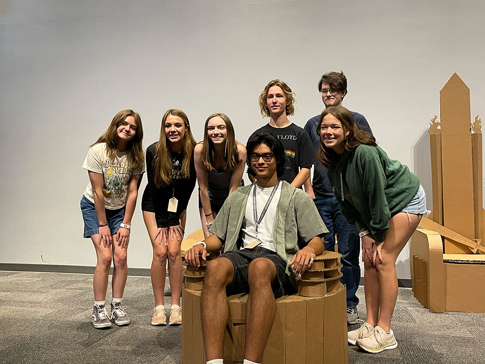 Group of campers pose around their cardboard chair project as one camper sits in it.