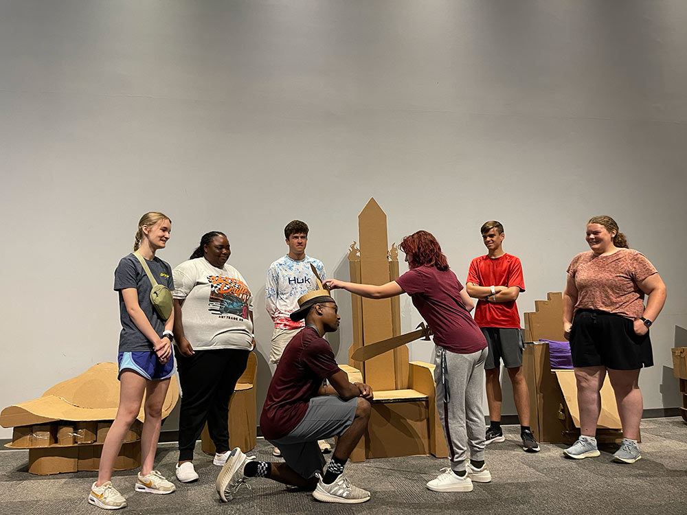Group of campers stand around their cardboard chair project as one camper kneels with a crown, and the other stands with a sword.