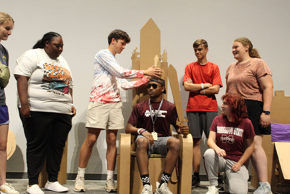 Group of campers stand around their cardboard chair project as one camper sits in it.