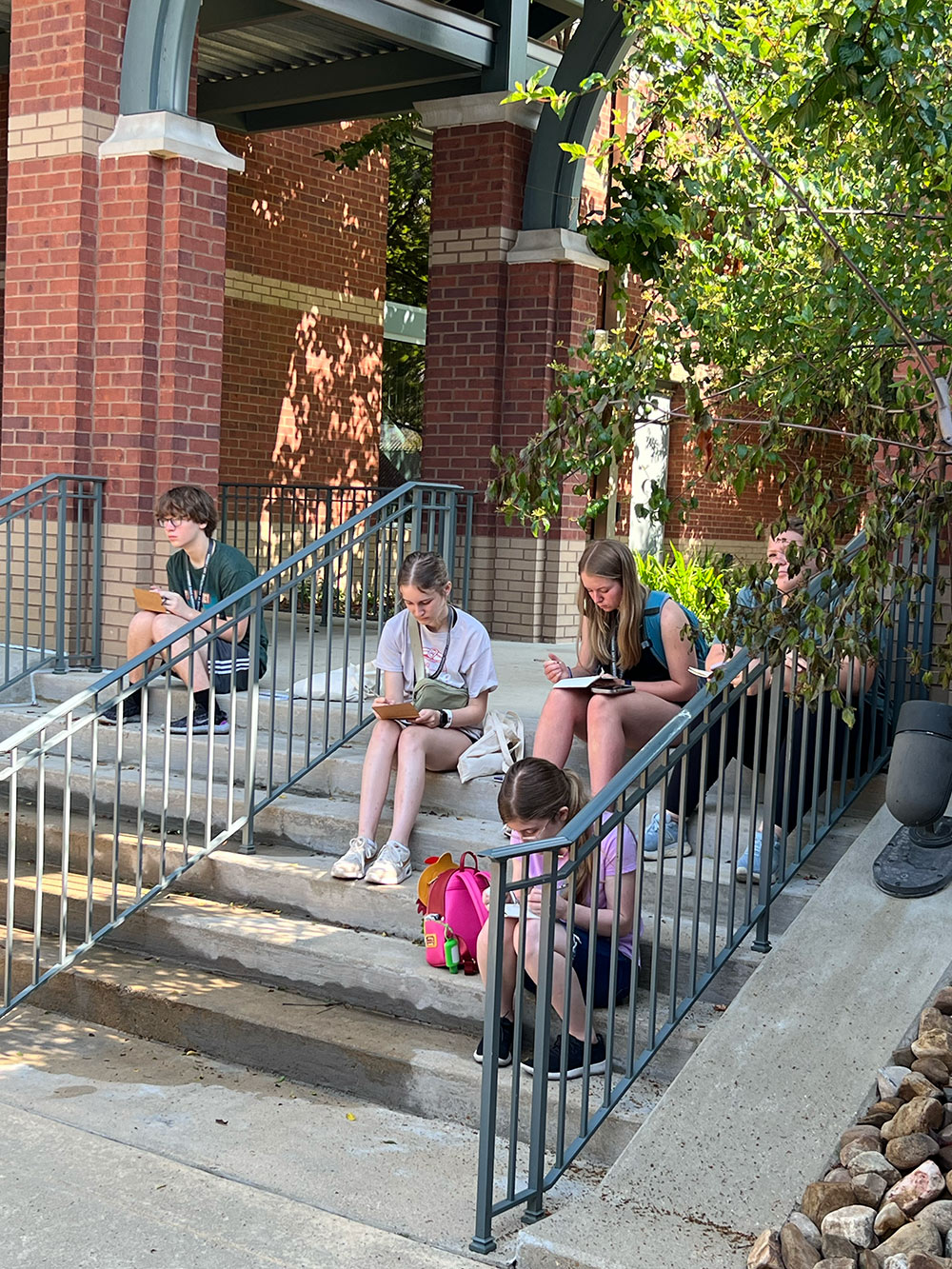Group of Design Discovery campers sitting on steps outside, sketching in their notebooks.
