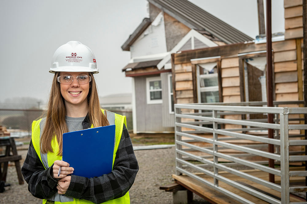 Mississippi State University Building Construction Science senior Jalyn Wallin stands with clipboard in hand (wearing hard hat and vest) in front of modular buildings built by first-year students.