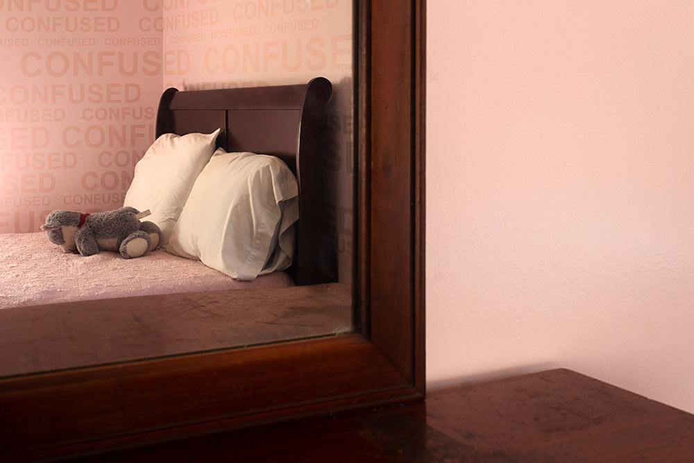 bed in pink room with gray stuffed animal on bed - "confused" written in repeat on wall behind bed