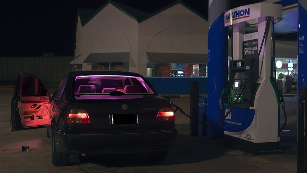 car with pink and orange glowing light and door open at gas pump; wallet on ground by open door
