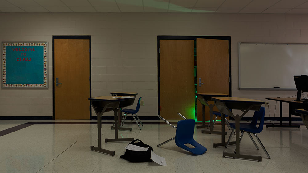 classroom with chair overturned, backpack on ground. Door is open with glow coming from crack