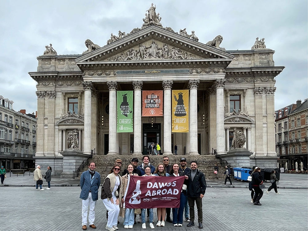 students on the Mississippi State University Building Construction Science Department's study abroad 2025 trip gather with MSU flag in front of building