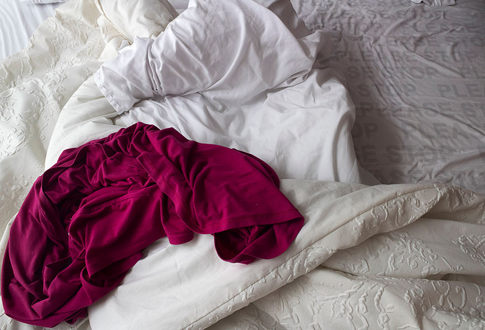 bed - unmade with white sheets and maroon/pink shirt on it - "please stop" on pillow