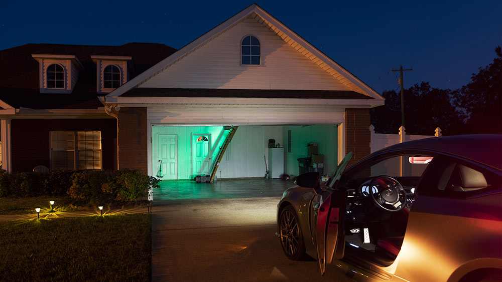 view from behind car that sits in driveway of house looking into open garage with attic stairs down and green glow
