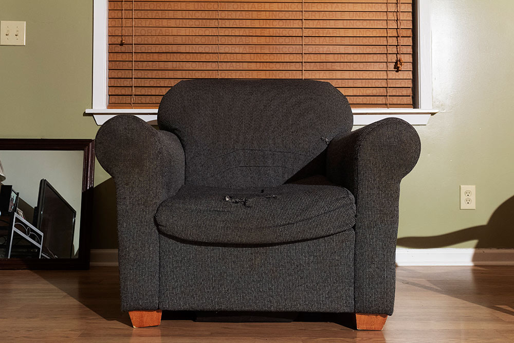 old, gray chair with "uncomfortable" written on wooden blinds behind