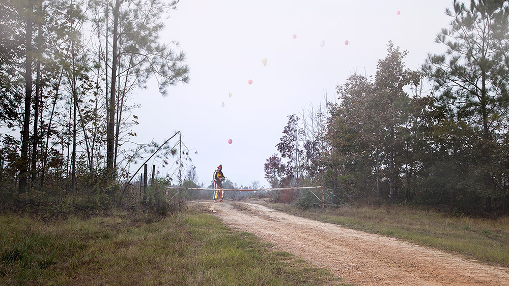 A clown stands along a gravel road in the woods with pink balloons flying above it.