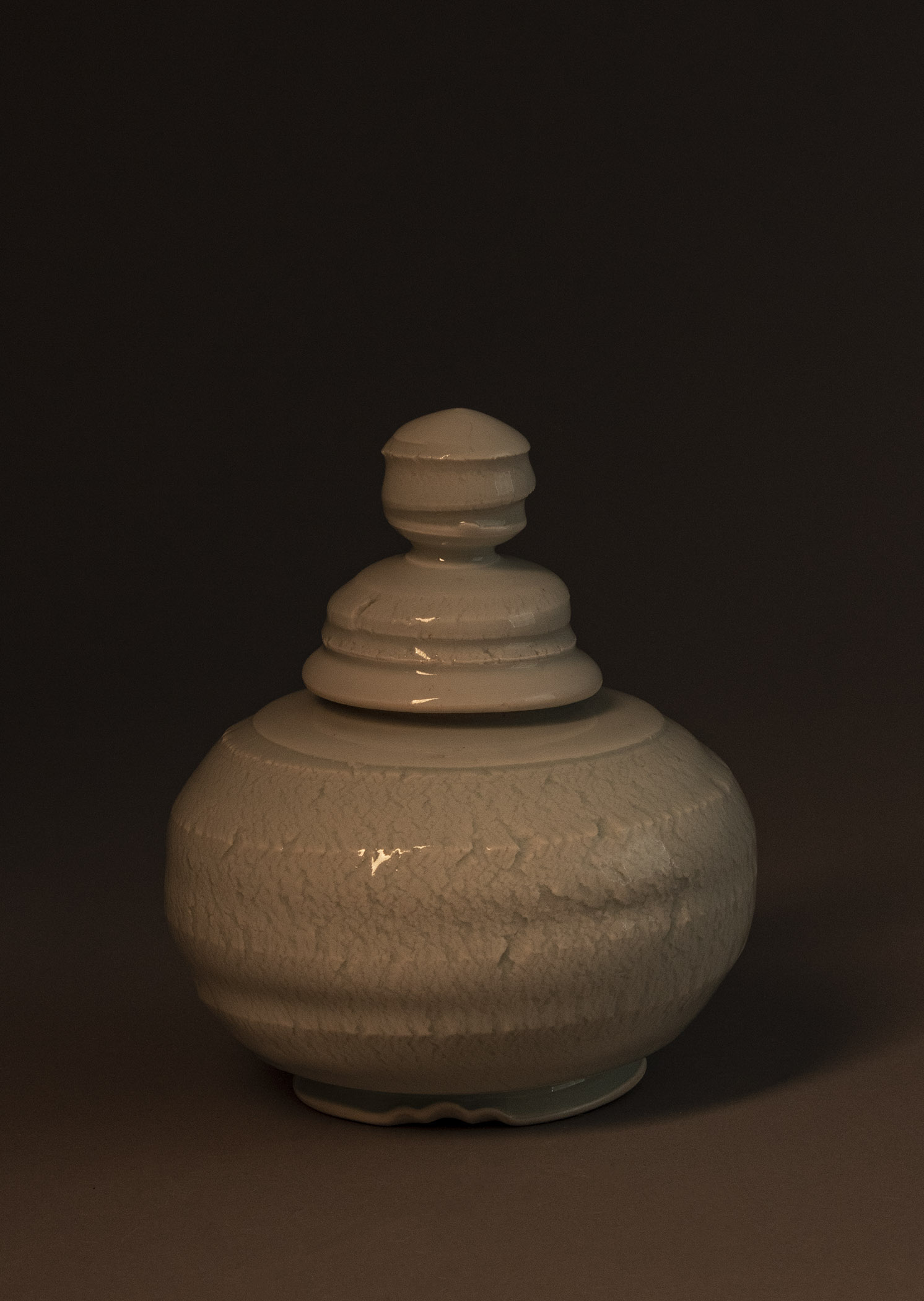 A lidded jar with a somewhat irregular orb-shaped body, a shallow ring-shaped foot that flares out at it’s base, and an irregular dome-shaped lid topped with a sort of twisting textured finial. The jar itself has the open texture of stretched clay. The lid and finial have ridges that spiral around their forms and show some stretching/cracking at their edges. The glaze is glassy.