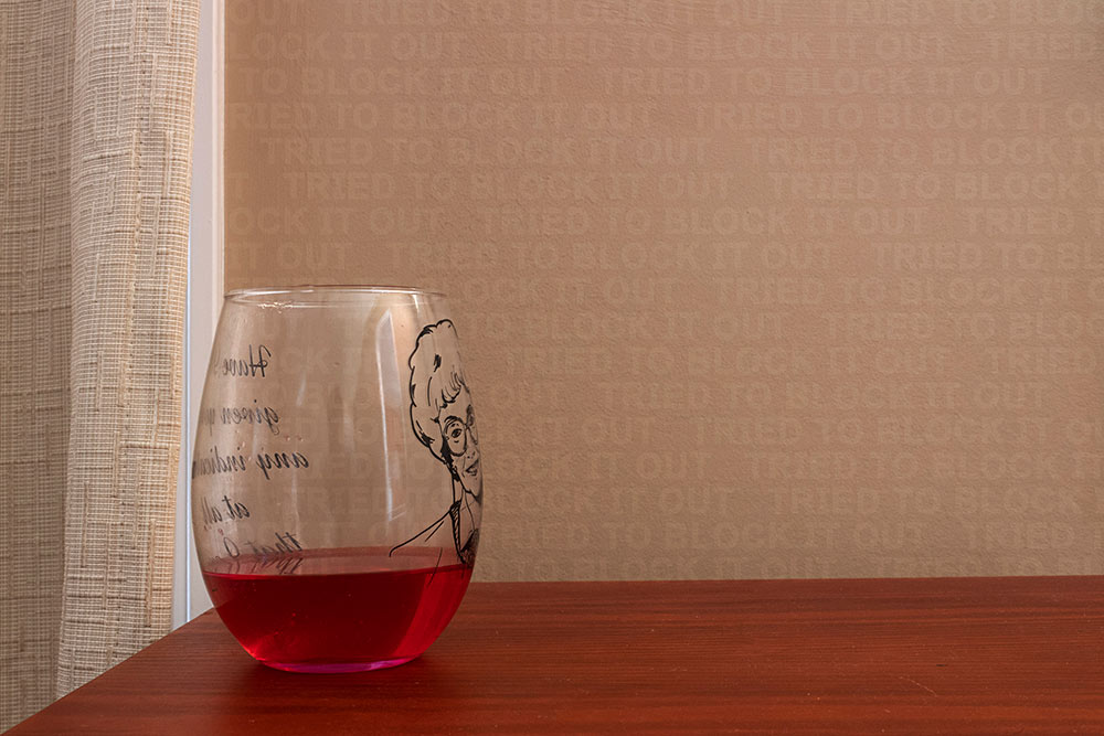 clear glass with red drink on wood table - hard to read letters in black (backwards) on left and woman's face on right of glass