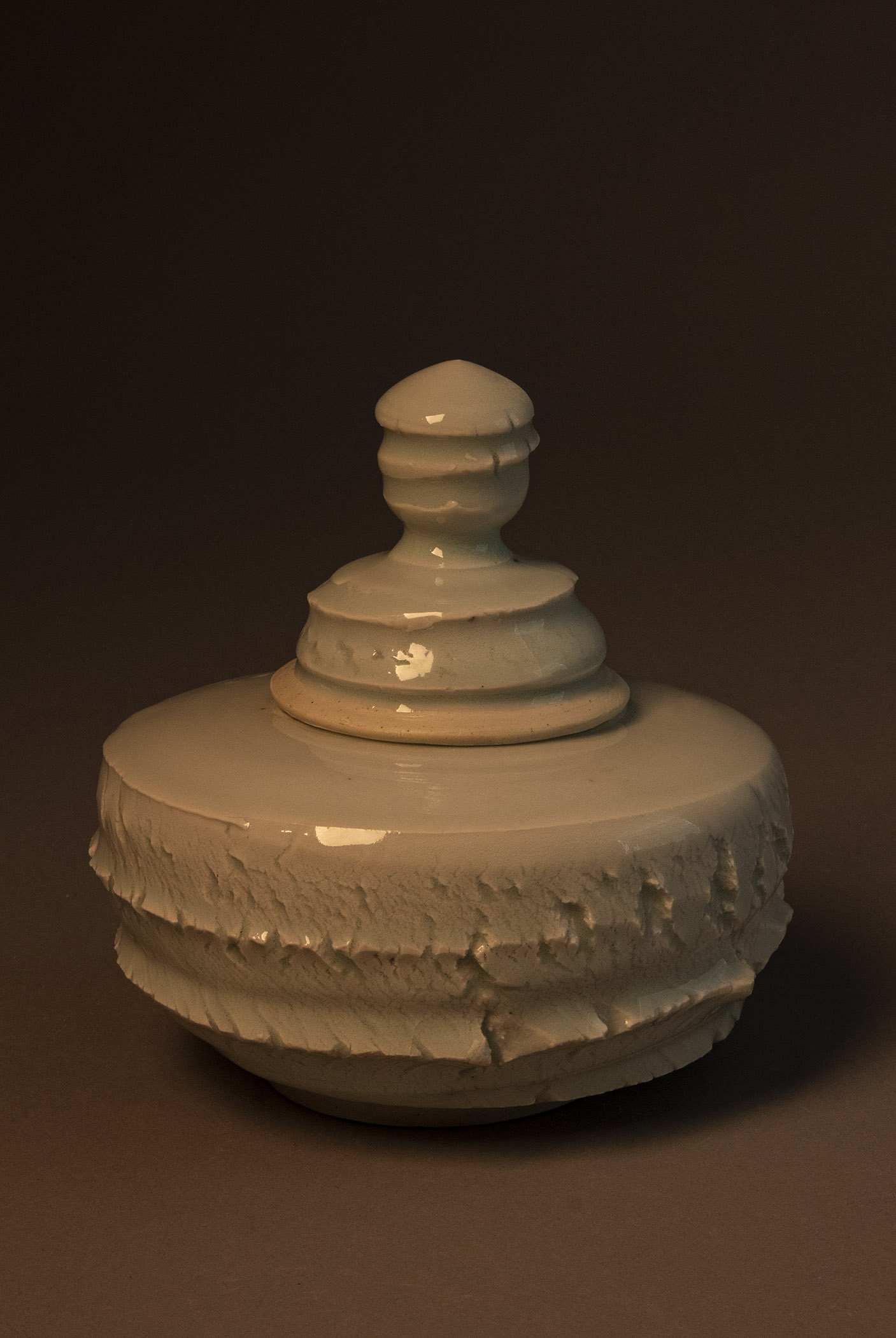 A lidded jar with a barrel-shaped body that is wider than it is tall. The body of the pot has nearly horizontal raised lines wrapping around it that are slightly abrasive to the touch. The lid is bell-shaped, but its profile is irregular due to the cutting into and stretching of the exterior surface. The lower rim of the lid is unglazed and smooth, while the upper portion and the knob toping it both have the texture of stretching and breaking clay. The glaze covering the piece (with the exception of the rim of the lid) is glassy.