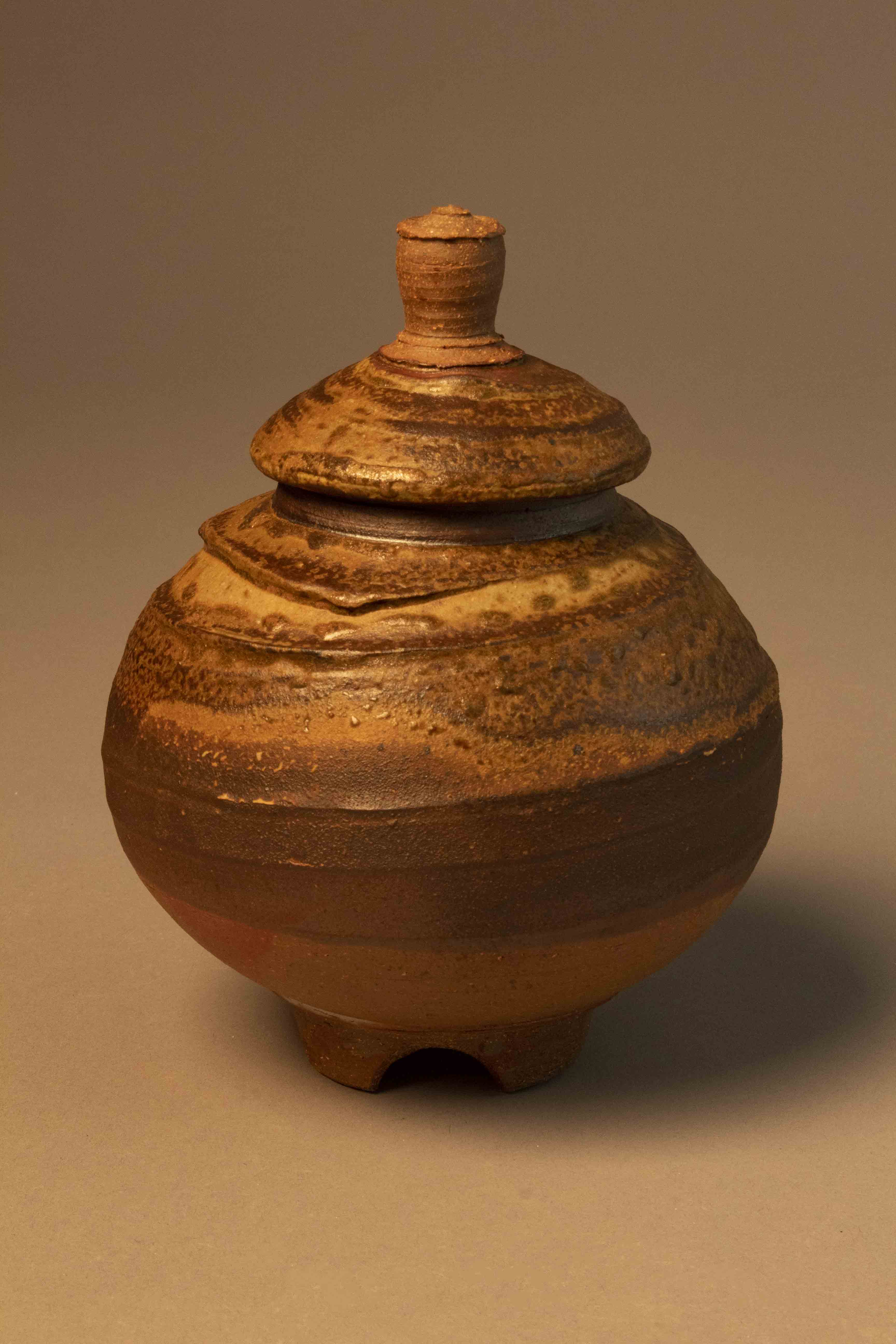 A jar with a pronounced rounded belly, a trimmed foot, and a lid that is shaped like a conical hat and is topped by a knob. The stoneware body is groggy and more course than the porcelain. The surface has been textured with slip so raised ridges spiral around the body of the jar as well as the exterior of the lid. Over this, a wood-ash based slip has been sprayed causing an uneven fluxing. The resulting surface has portions that feel drier while others may be smooth to the touch. The foot and the funnel-shaped knob are unglazed.