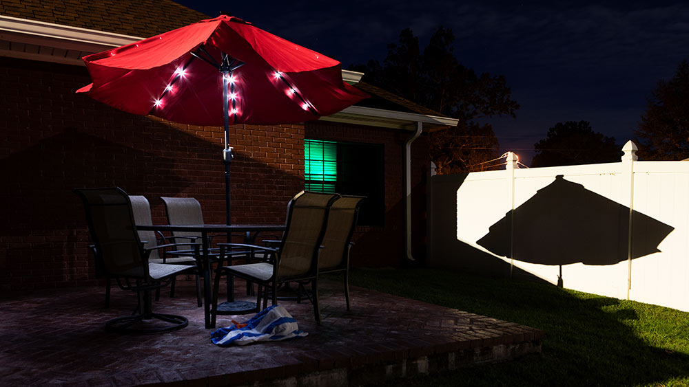 patio with umbrella. lights on umbrella and shadow of umbrella on white fence at right