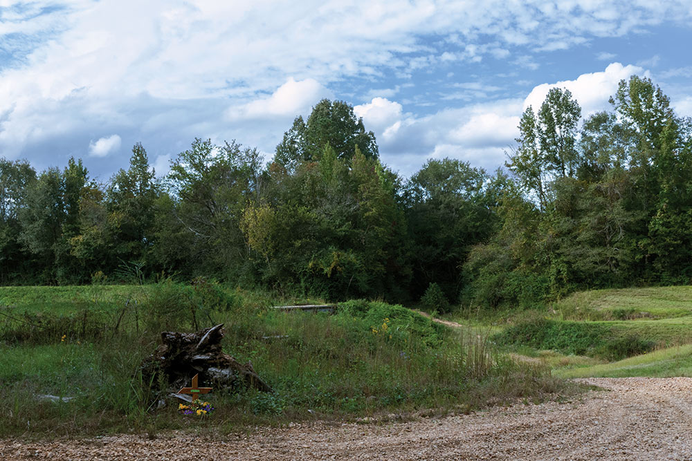 gravel road in woods - wood cross with flowers at center of photo next to road and wood pile