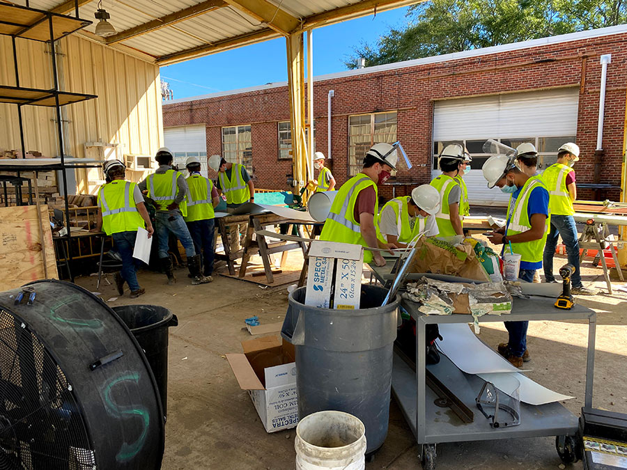 lots of architecture and building construction science students wearing construction PPE and masks work on parts of their modular building in the outside space behind Howell Hall