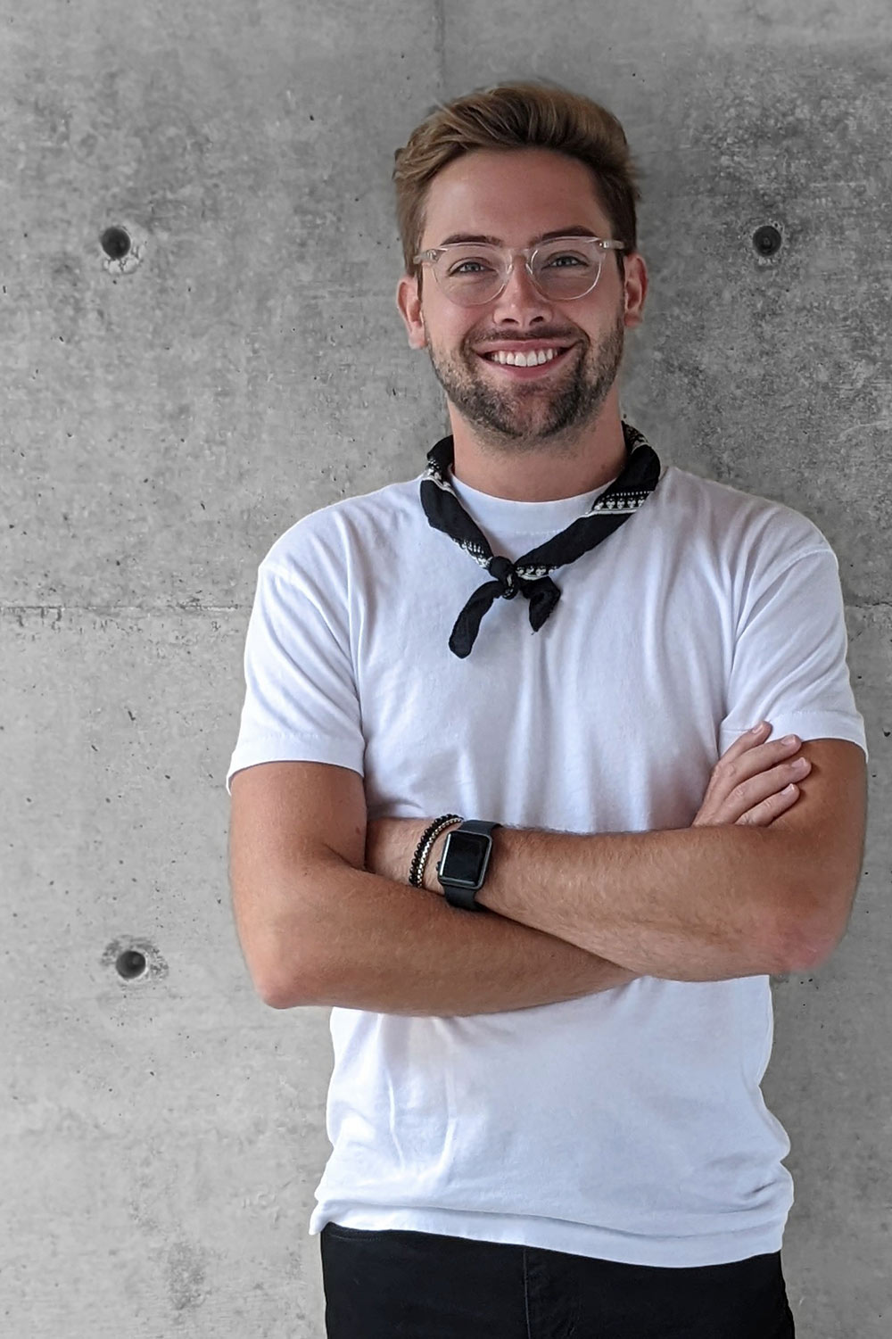 Zachary Henry-Litchliter posing with his arms crossed and smiling at the camera in front of a concrete wall wearing a white shirt and black bandana around his neck..